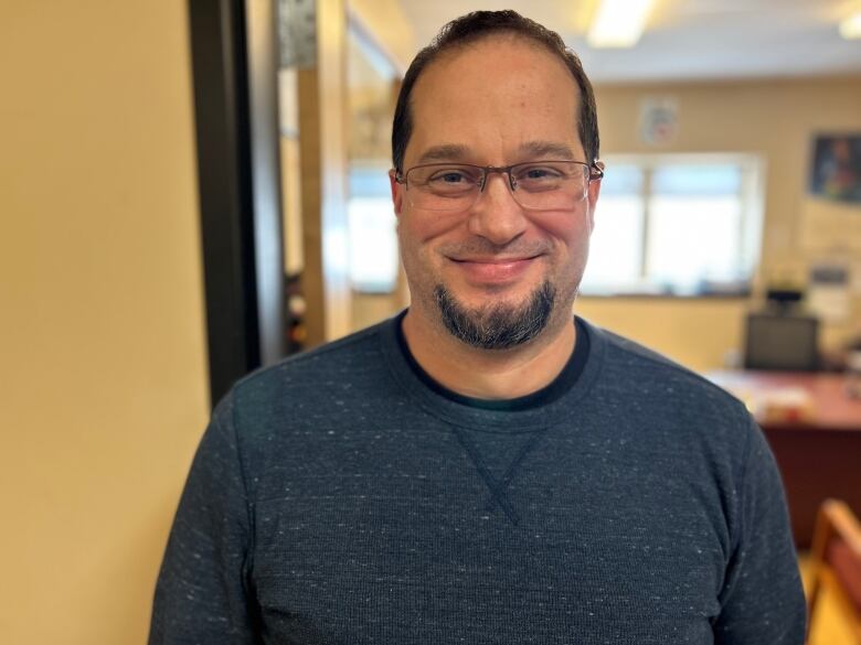 Man with glasses and wearing blue sweater stands in front of bulletin board.