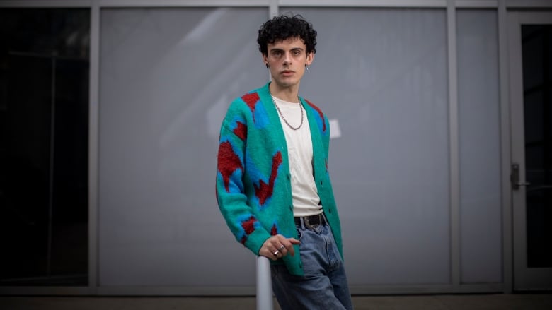 A man with curly black hair and a prominent nose looks directly at the camera. He's wearing a green, red and blue sweater, two different earrings and a silver necklace.