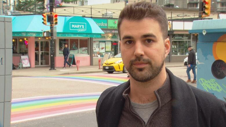 A man stands on a corner of Vancouver's Davie Village. He has short, black hair, a goatee and is wearing a wool jacket. The crosswalks are painted with rainbows.