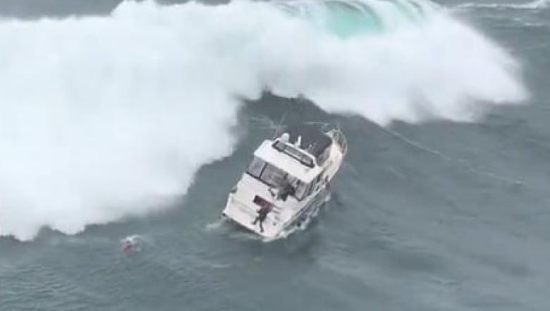 A huge wave is about to crash into a yacht on the open ocean as one man grips the back of the vessel and another swims toward it.