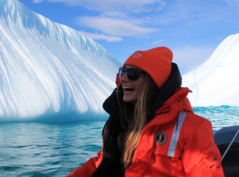 A young woman sits in a boat, looking off-camera. She has a big smile, with her mouth open and her teeth showing. She's wearing a life vest and sunglasses. In the background, there are several icebergs.