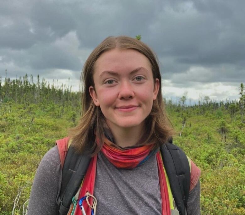 A young woman smiles for the camera. She is carrying a backpack. In the background, there are trees, bushes and grass.