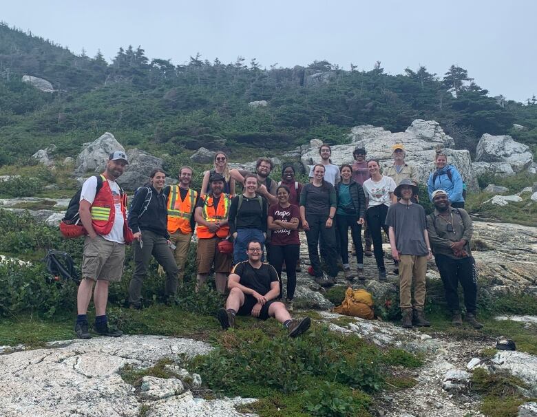 A group of 19 people stand together, posing for a photo. Some of them wear high-visibility vests and a few backpacks lie on the ground. The group is standing on a rock, with trees in the background.