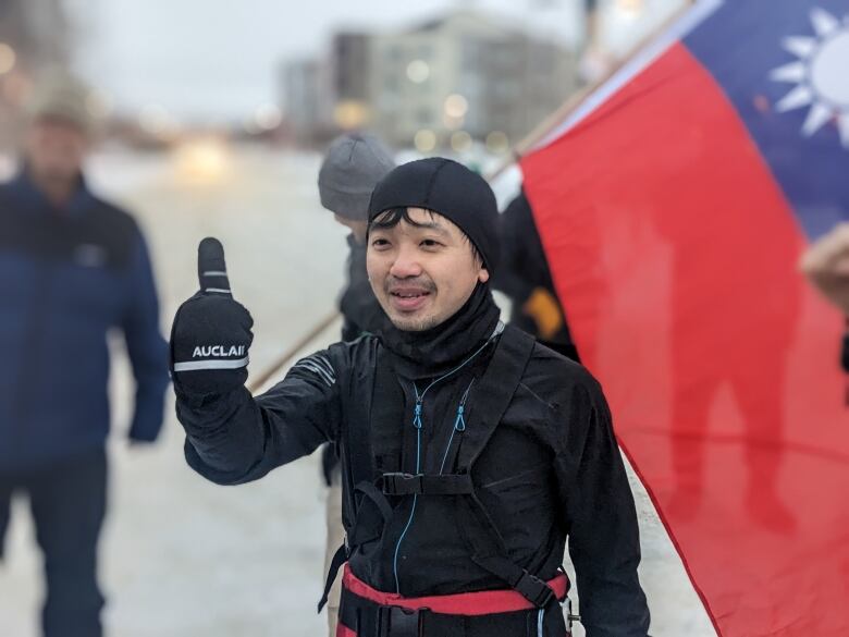 A man wearing a black beanie is giving a thumbs up, standing in front of a Taiwanese flag. 