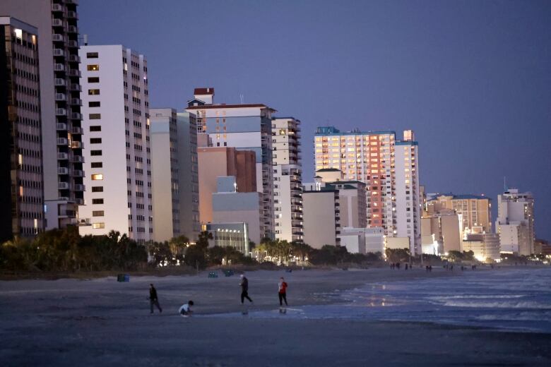 People walk on a beach at dusk with buildings in the background. 