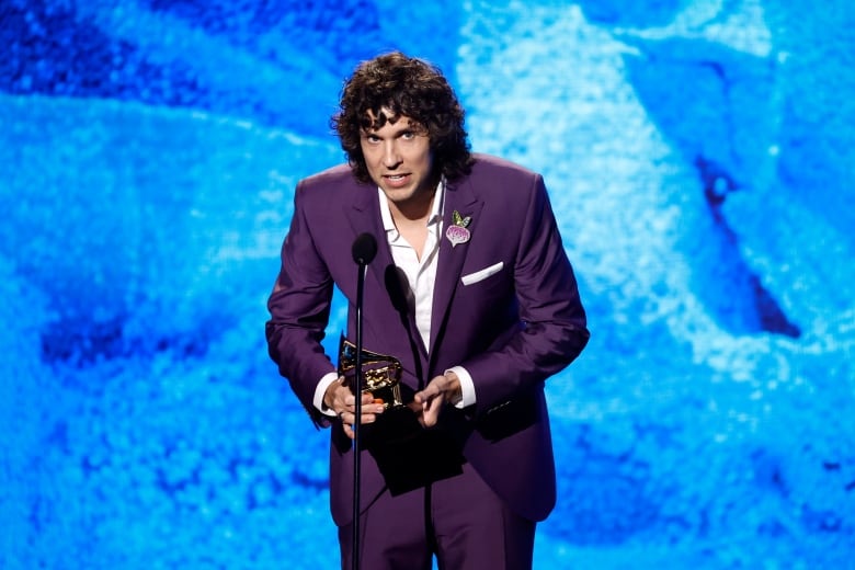 A man in a purple suit, holding an award, stoops to speak into a microphone. 