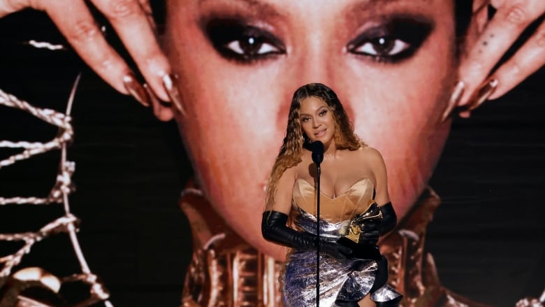 A woman, wearing a gown and holding a trophy, speaks at a microphone on stage. 