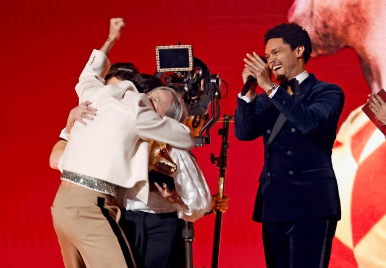 A man facing away from the camera hugs a smiling elderly woman holding a trophy. To their right a man smiles and claps. 