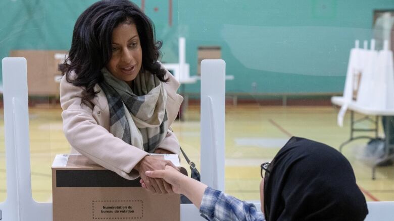 A woman shaking hands with another woman sitting behind a desk with a ballot box on it. 