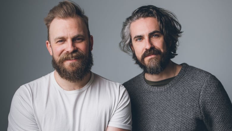 Two men smile at the camera. One, at left, short, spiked blonde hair and a beard. The one at right long, wavy, grey and white hair and a beard. 