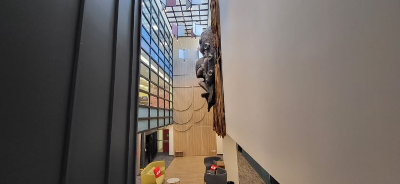 A series of coloured windows to the left. A giant piece of the art hangs on the wall to the right (an Indigenous mask) Behind them is a CBC logo carved into a wooden wall. Below are a yellow couch and black chairs with CBC branded red pillows