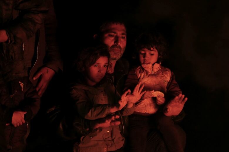 A man and two children sit in the red glow of a fire outdoors. 