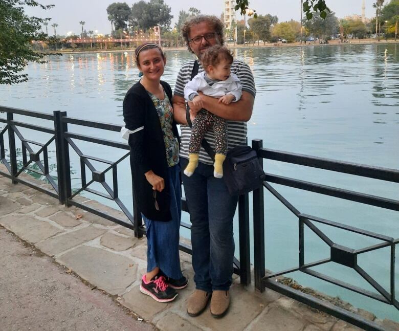 A man and a woman stand by the water and smile. The man is holding a small child. 