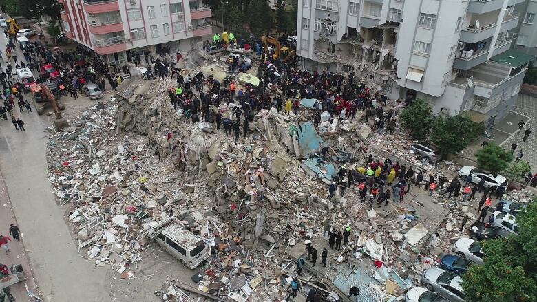 People work atop, and gather around, a huge mound of rubble between heavily damaged buildings.