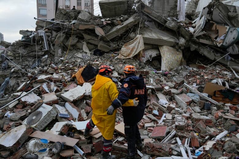 Two rescue workers stand with their backs to the camera on top of a pile of rubble, their arms around each other.