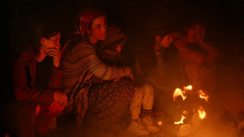 A woman and four young girls sit around a fire with exhausted expressions on their faces. 