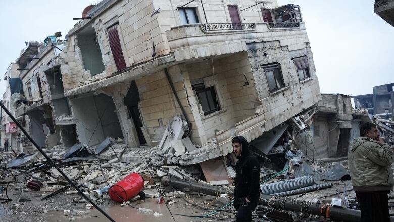 A man stands in front of a damaged concrete building that sits slanted off its base.