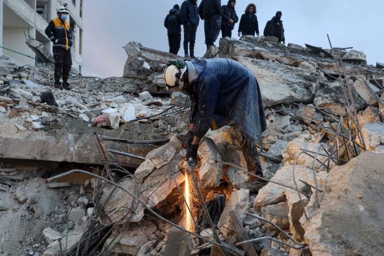 A man with a white helmet cuts through concrete as other people stand above, on a pile of rubble, watching. 