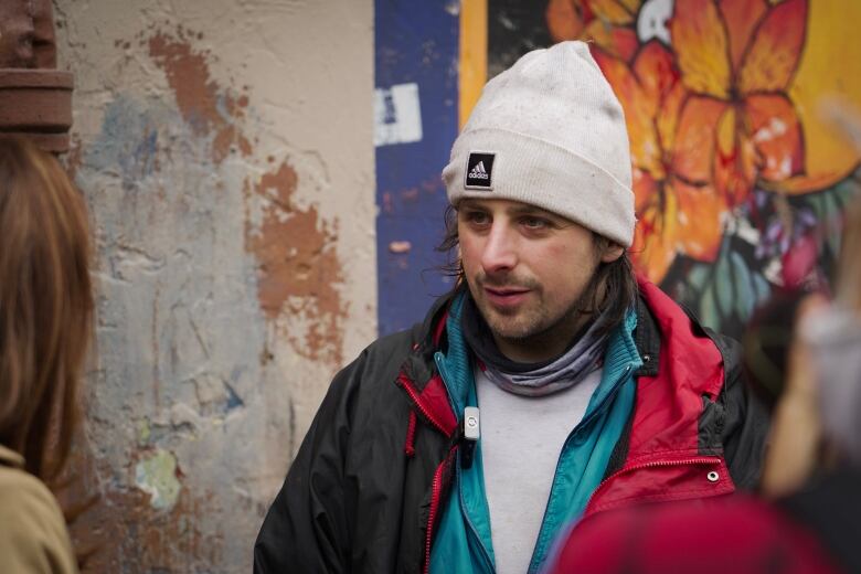 A man in an Adidas toque and numerous jackets looks at a reporter just out of frame.