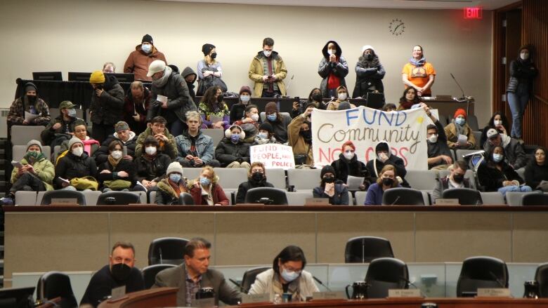 Protestors hold signs in a gallery seating area while three councillors sit around the table.