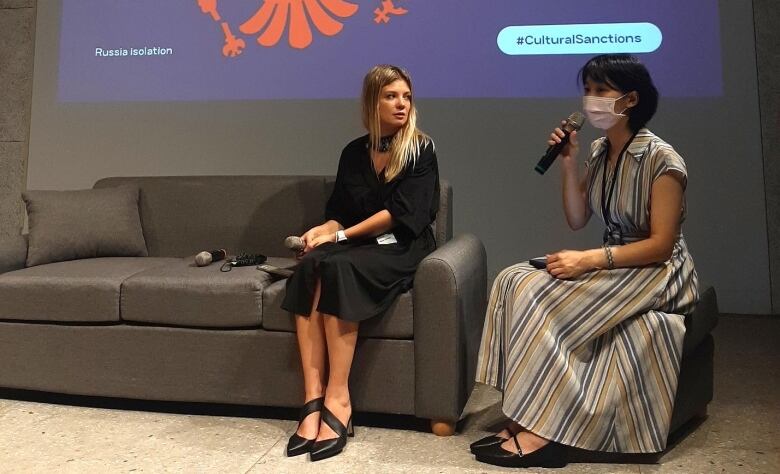 Woman with long blonde hair and wearing black dress sits on sofa next to woman with short black hair and wearing a face mask and dress, and holding a microphone, under a screen with text Impose Cultural Sanctions On the Russian Federation.