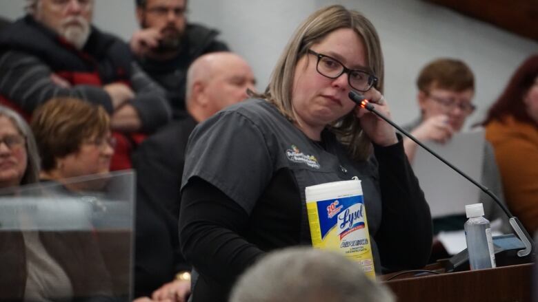 A woman at a microphone podium looking at someone speaking to them out of frame. 