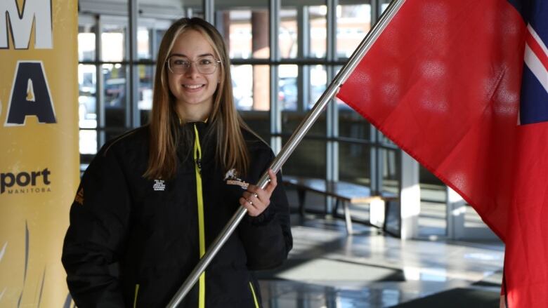 A smiling girl wearing glasses and a Team Manitoba zip-up holds a Manitoba flag.