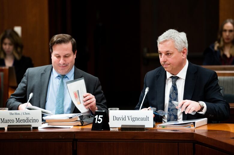 Two men in suits, with binders of papers in front of them, sit at a table.