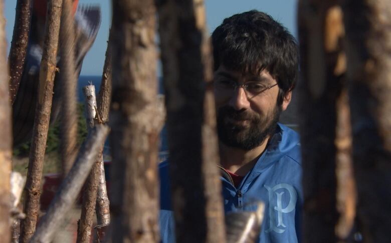 A bearded man with glasses is seen through a series of large sticks used to make a fence. 