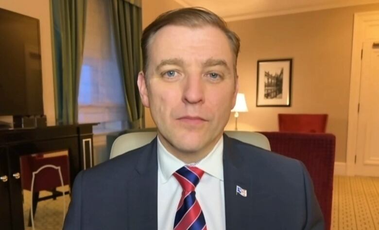 A man wearing a black suit and white shirt sitting in a hotel room. 