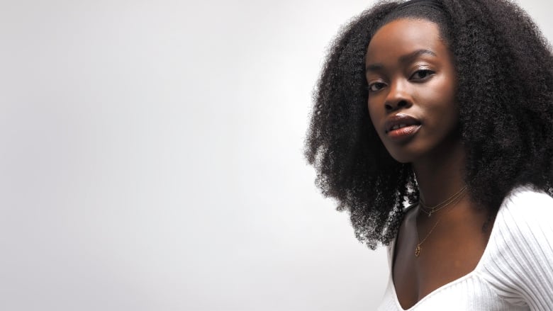 A young Black woman in a white knit shirt looks at the camera.