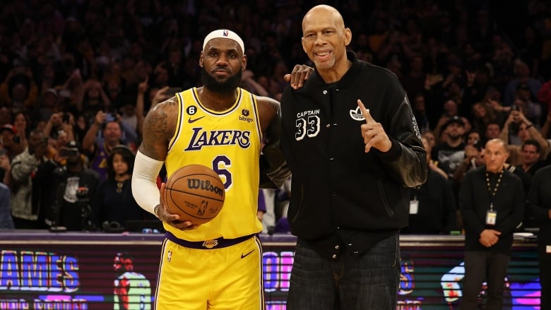 A basketball player wearing a yellow Los Angeles Lakers uniform holds a ball as he poses to a picture with taller man wearing black clothes. 