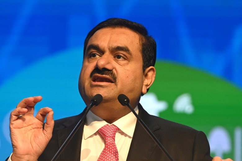 A man with a mustache, wearing a dark suit and red tie, gestures as he speaks before a microphone.