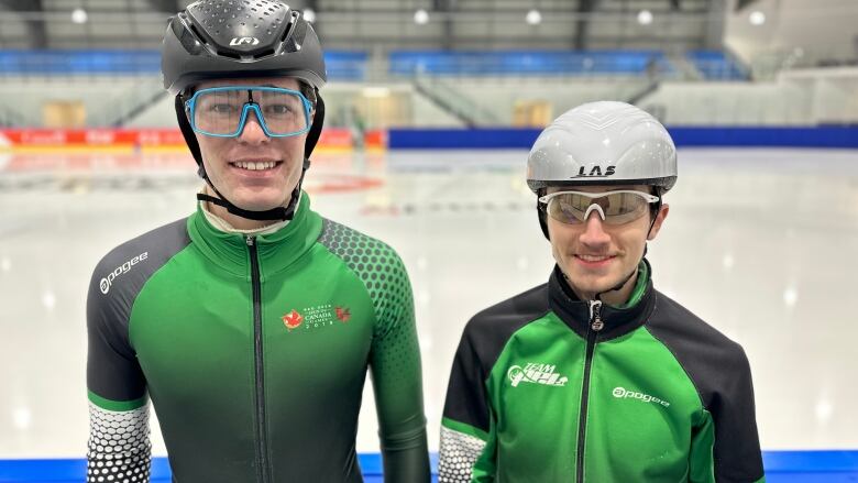 Two speed skaters wearing goggles and helmets stand with the ice in the background. 