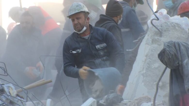 A man wearing a blue jumpsuit and a hard hat uses a blue plastic bucket to move rock, as others work behind him. In the foreground are the remains of a levelled building.