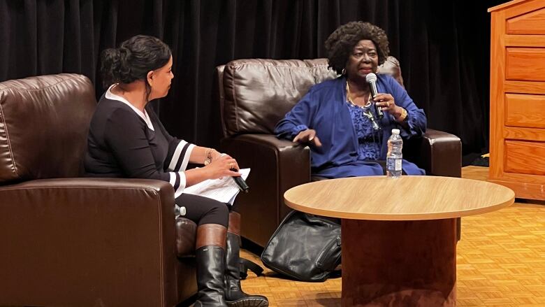 A Black woman sits in an armchair on a stage, speaking into a microphone, with another woman sitting in a chair beside her listening.