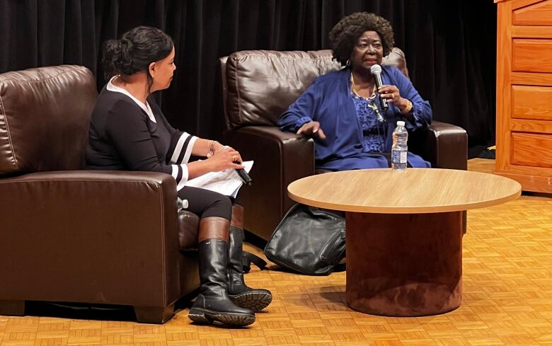 A Black woman sits in an armchair on a stage, speaking into a microphone, with another woman sitting in a chair beside her listening.