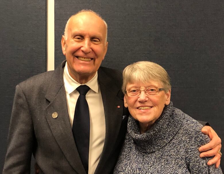 Older grey haired man in suit smiles with his arm around an older grey haired woman in sweater, also smiling