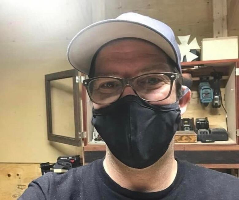 A man in a baseball cap, a black face mask and glasses poses for a selfie.