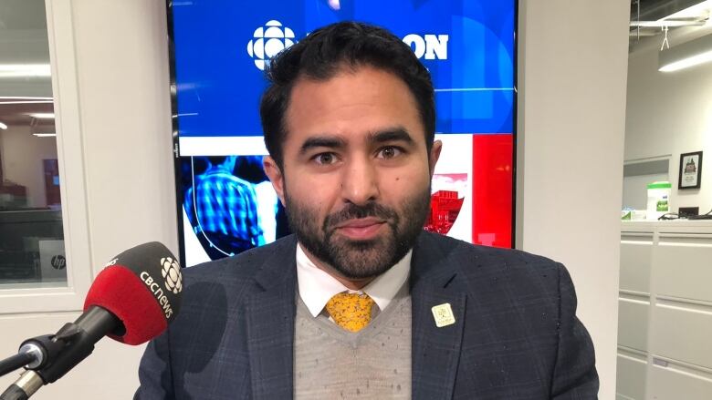 London's first Muslim Community Liaison Advisor Ahmer Khan sits at a microphone at the CBC London studio wearing a blazer, shirt, tie and sweater.