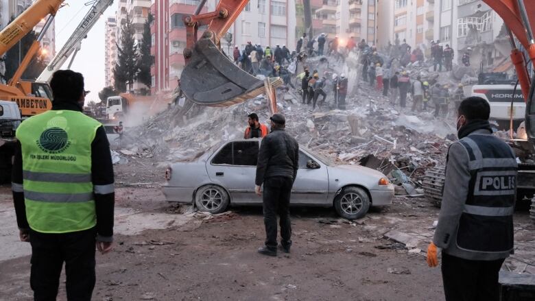 Several men stand with their backs to the camera, at various distances away, in front of a large pile of rubble and a huge orange digger.