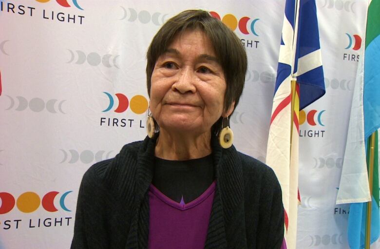 A woman wearing a black sweater and purple shirt standing in front of a backdrop which reads First Light. 