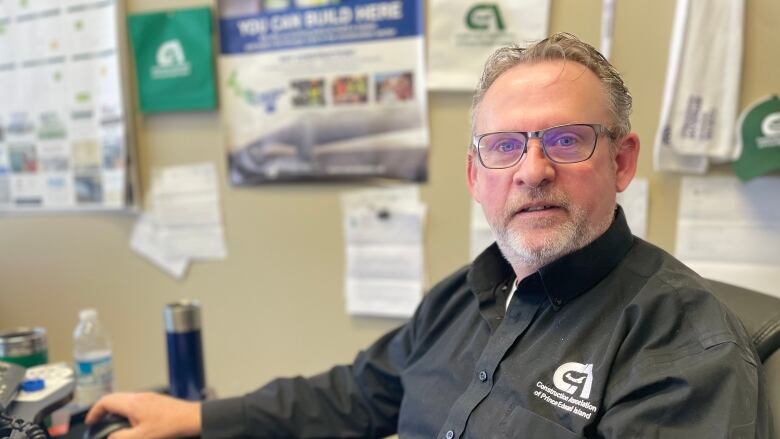 Man with short grey hair and glasses sits at computer wearing a dark shirt with branded logo for the Construction Association of P.E.I.