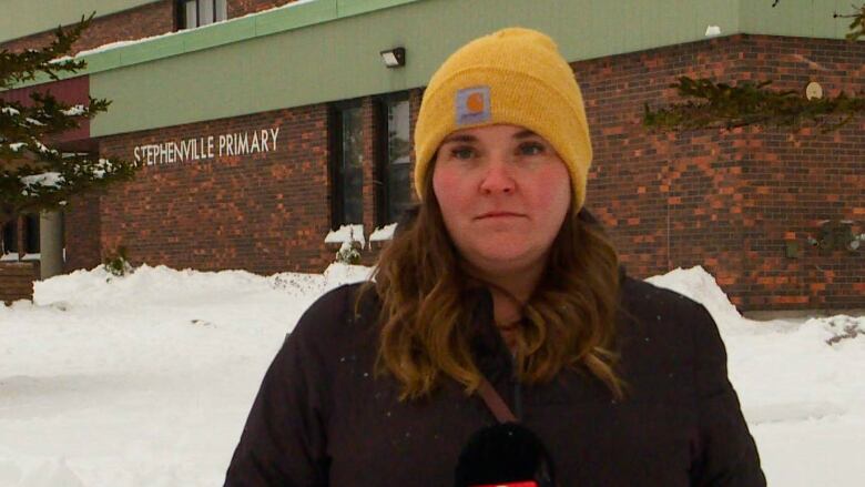 A woman with long, brown hair and a yellow hat stands in front of a brick school