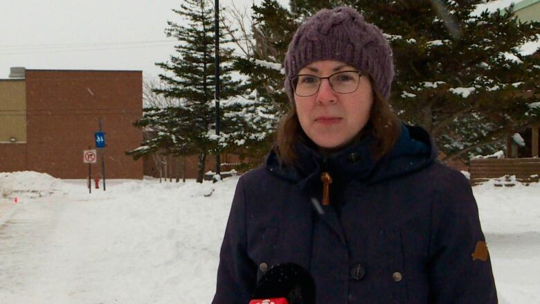 A woman with medium-length brown hair and a purple hat stands in front of a brick school