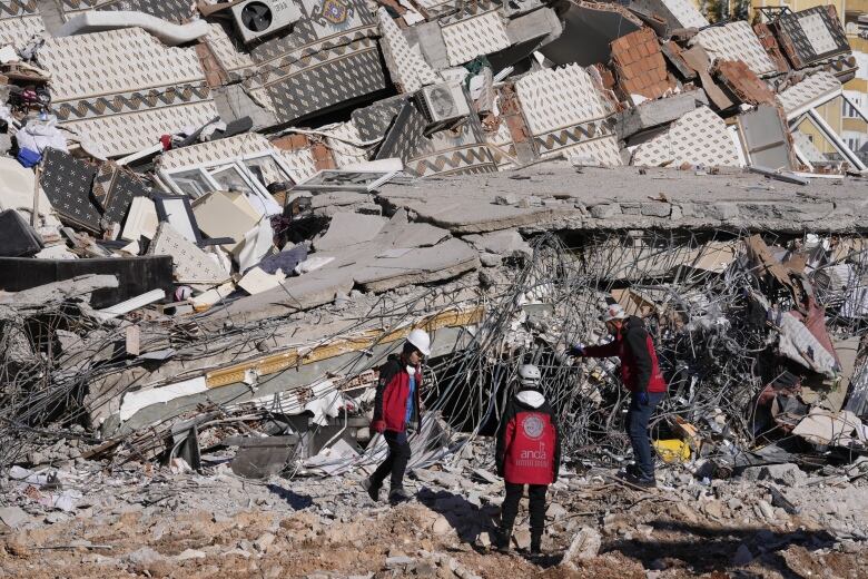 Three people wearing black and red jackets and white hard hats stand next to a massive pile of rubble, including large chunks of concrete and twisted metal.