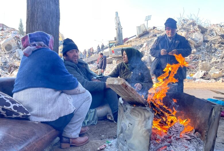 Two women and two men huddle around the open flame of a fire, seated outside. Behind them is the wreckage of collapsed buildings.