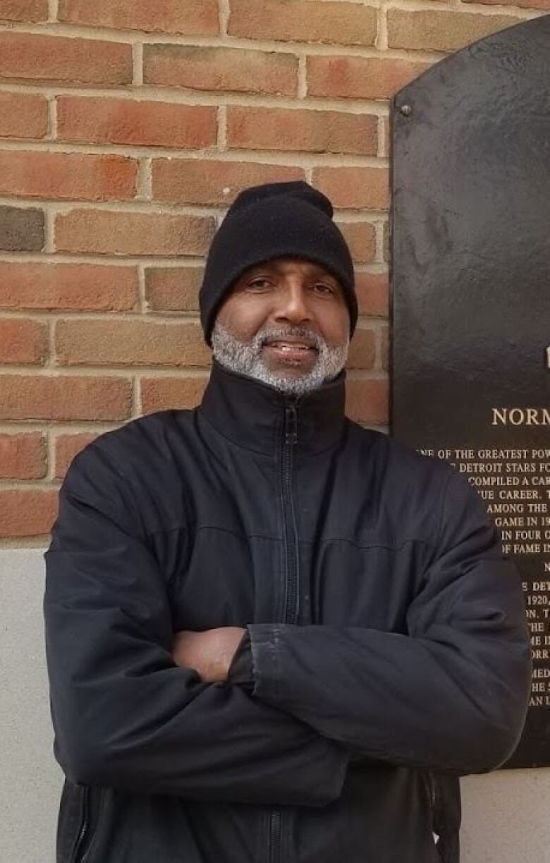 A man wearing a black jacket and hat standing next to a plaque honouring a baseball player