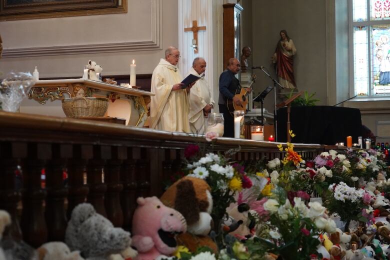 A priest and other church officials speak on an altar lined with stuffed animals and bouquets of flowers.