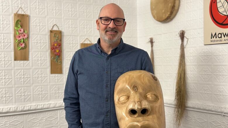 A man wearing a blue shirt stands holding a large, wooden mask.